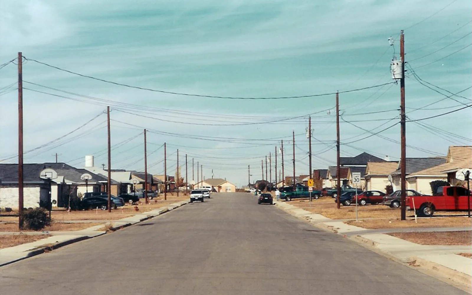 A quiet suburban street lined with houses, mature trees, and sidewalks under a blue sky
